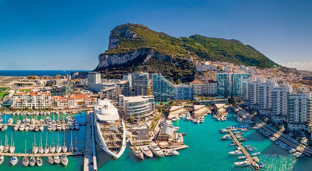 Aerial view of a Mediterranean marina with luxury yachts, turquoise water, and a hillside city in the background.